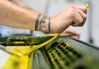 Nahaufnahme einer Hand die gelbe Druckfarbe in die Walze einer Druckmaschine gibt.